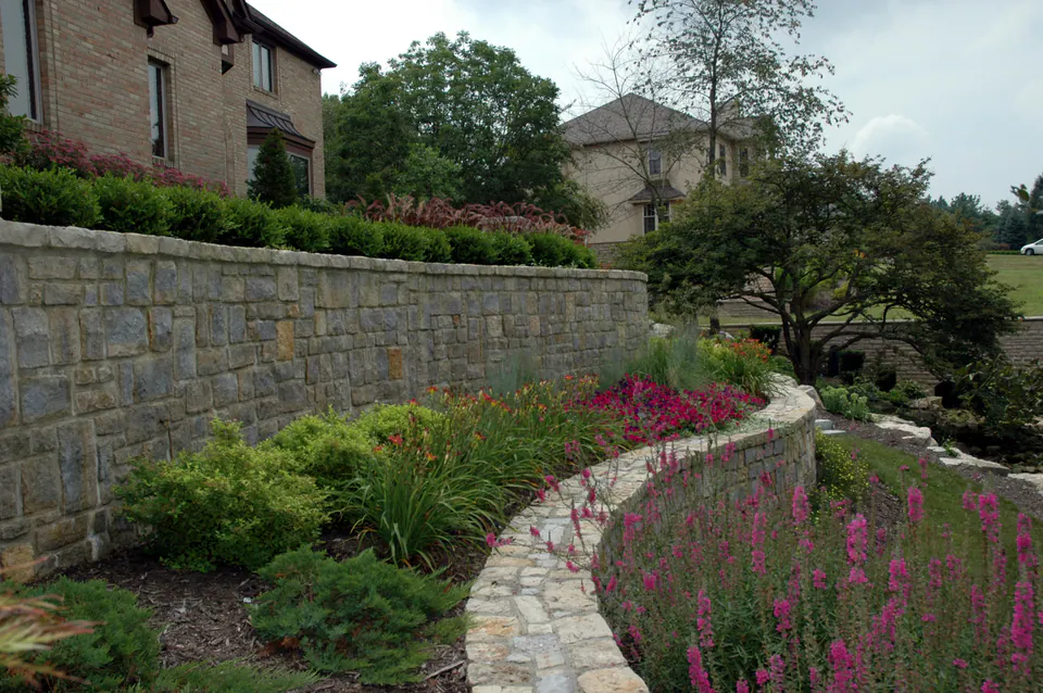 Tall curved stone retaining wall above a bed of pink perennials and evergreens beside a brick house